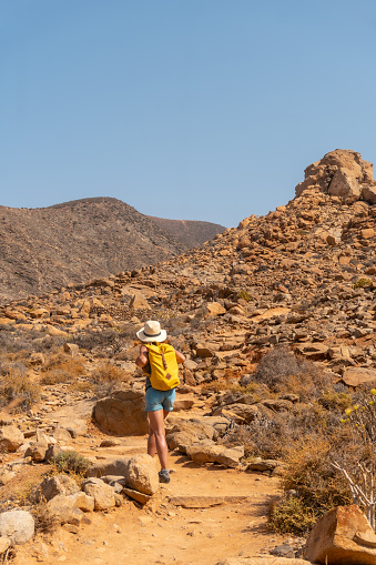 A vertical shot of a girl hiker with a yellow backpack on the Mirador de la Penitas trail in the Penitas canyon, Fuerteventura, Canary Islands, Spain