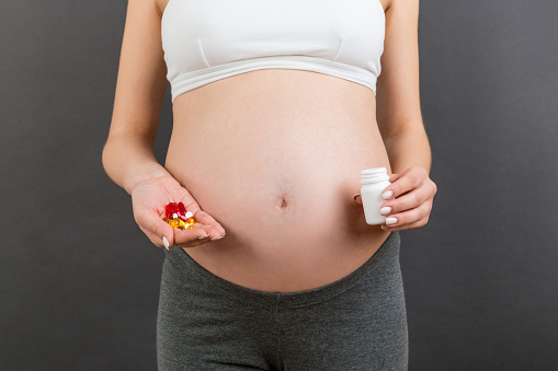 Close up of pregnant woman holding a bottle and a pile of pills in hands at colorful background with copy space. Healthcare concept.