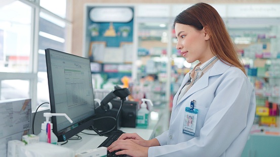 Professional asian woman pharmacist using a computer to research medicines and recheck stock of products in the pharmacy