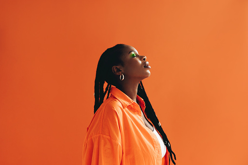 Fashionable african woman with dreadlocks standing in a studio with her eyes closed. Confident black woman wearing piercing jewellery and makeup. Female hipster standing against an orange background.