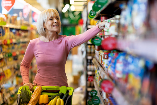 Positive attractive elderly woman with supermarket trolley standing by shelf with food and smiling, choosing goods at store, happy with assortment, copy space. Quality food concept
