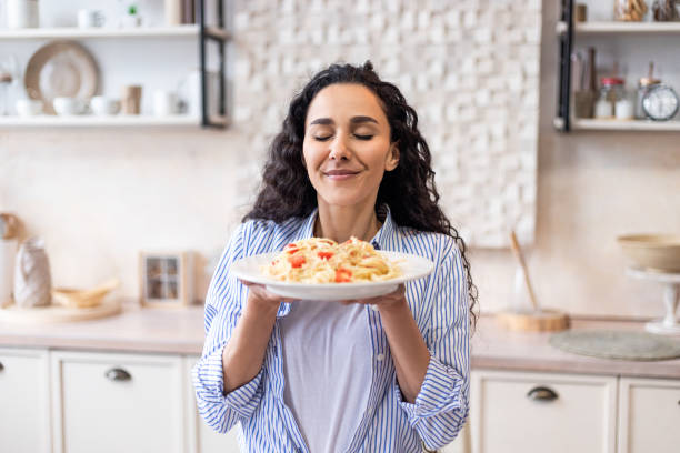 señora feliz degustando espaguetis caseros mientras almuerza, disfrutando del olor con los ojos cerrados, sentada en la cocina - tasting women eating expressing positivity fotografías e imágenes de stock