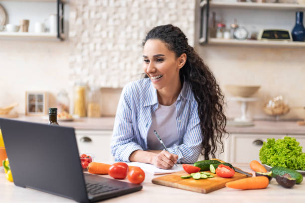 istock Writing down new recipe. Happy latin woman taking notes in copybook while watching cooking webinar on laptop 1466358371