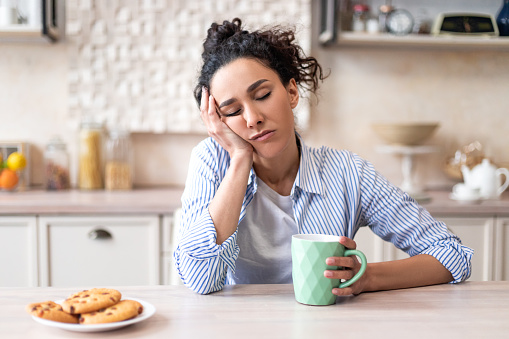 Sleepy young woman sitting at dining table in kitchen with closed eyes, holding cup while having breakfast. Early wake up, insomnia, lack of sleep