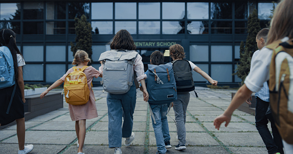 Diverse children going school with backpacks back view. Cheerful smiling teachers meet welcoming teen students at open door. Excited elementary age pupils entering modern building. Study concept.