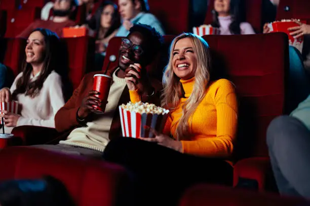 A cheerful multiethnic young adult couple are in a movie theater having fun while watching a movie, enjoying popcorn and soda.