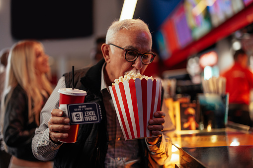 A senior Caucasian man is in a movie theater carrying popcorn, soda and tickets in his hands on his way to the movie hall.