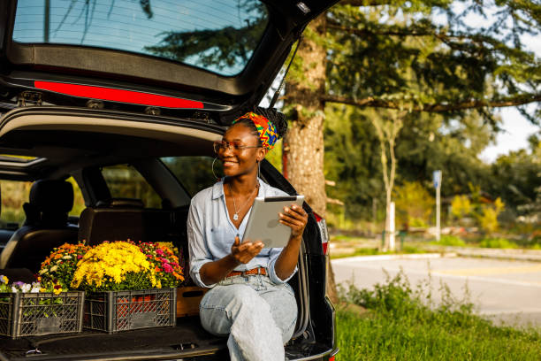 Young woman sitting in open car trunk, using digital tablet