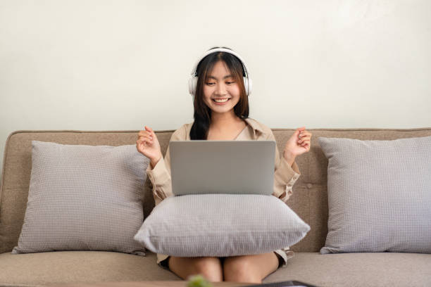 istock Female entrepreneur is sitting on big comfortable sofa and putting laptop on pillow while wearing headphone 1466392700