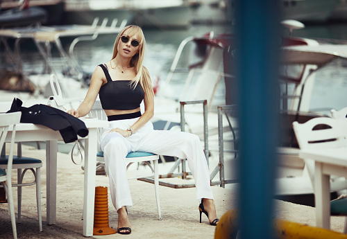 Beautiful and noble woman sitting on a white chair at a yacht marina.