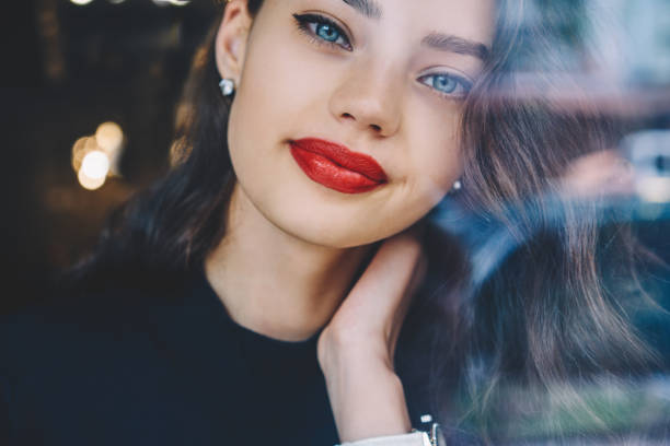 View through glass of crop attractive young lady wearing black clothes and white wristwatch touching neck while looking at camera
