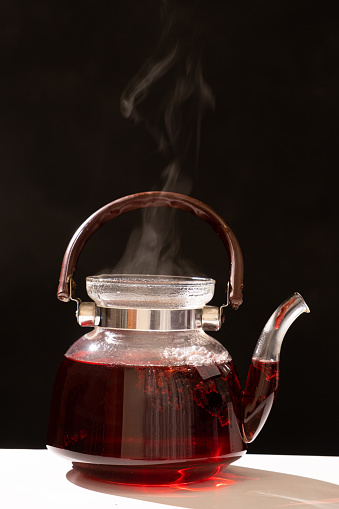 Red hibiscus tea from the petals of a Sudanese rose in a glass teapot, steam from a teapot on a black background.