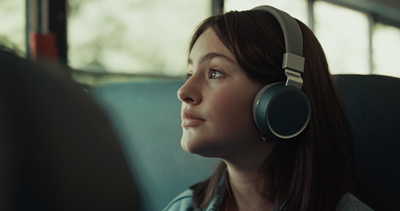 Thoughtful girl teenager traveling bus alone with headphones close up. Cute brunette student listening music with headset looking window. Pretty schoolgirl passenger enjoying daily ride to school.