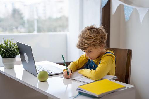 Caucasian child schoolboy or girl studying at home using laptop remote education. Doing homework, writing exercise book. Copy space