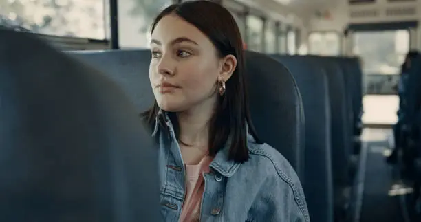Focused teenage girl sitting in empty schoolbus alone. Cute tensed student looking window on daily ride. Unhappy dark-haired schoolgirl passenger feel lonely riding to school. Teen lifestyle concept