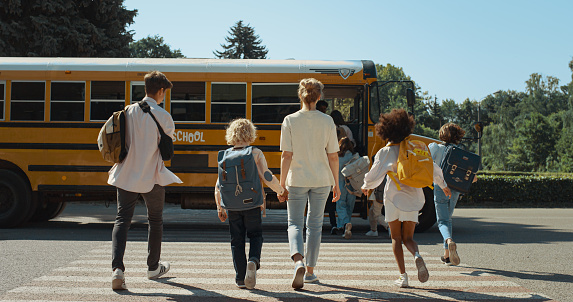 Mother hold hand schoolboy walking to yellow schoolbus. Energetic teenage pupils walking crossway. Young mom accompany curly son to school sunny morning. Teens crowd running boarding academic shuttle.