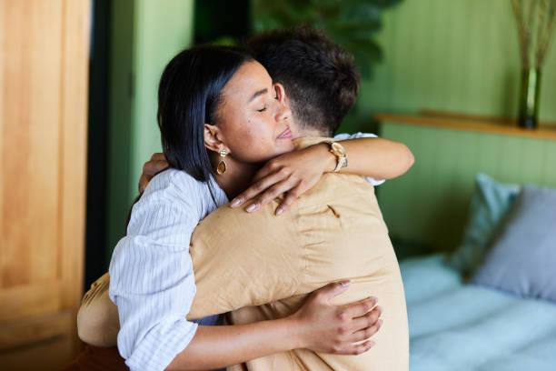 Loving woman hugging her upset husband in their bedroom at home Young woman consoling her husband with a hug while sitting together on their bed at home embracing stock pictures, royalty-free photos & images