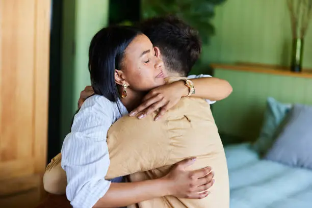 Young woman consoling her husband with a hug while sitting together on their bed at home