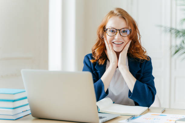 istock Positive redhead executive manager searches information on internet website, touches both cheeks, writes notes in notepad, uses modern laptop computer, dressed in formal clothes, checks organiser. 1466411992