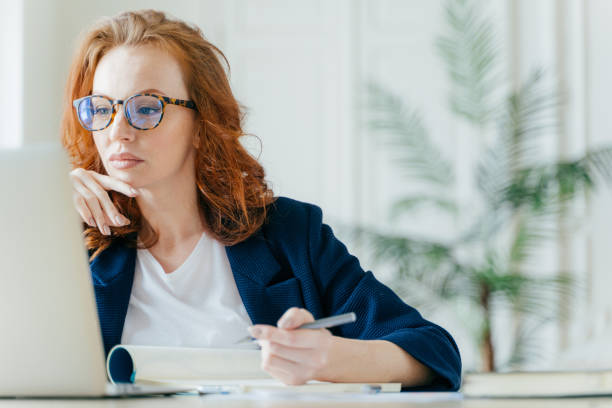 istock Serious experienced female coach makes records in notepad, focused at screen of laptop computer, has long red hair, wears transparent glasses, watches webinar online. Business and job concept 1466412013
