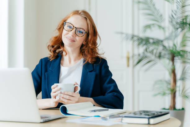 istock Pleased female teacher makes research work on laptop computer, writes down information in notepad, enjoys drinking coffee, has happy smile on face, sits in spacious room at desktop plant in background 1466412029