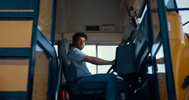Professional man driver sitting school bus cabin alone. Young chauffeur wearing uniform unlock entryway outside view. Serious worker look in camera opening vehicle door. Transportation concept.