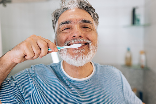 Cheerful smiling mature Caucasian man in pajamas washing teeth close up