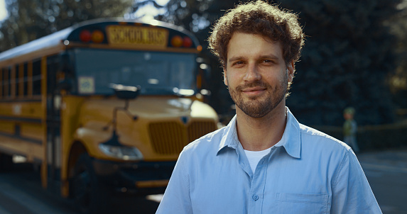 Smiling man driver posing near yellow academic vehicle alone close up. Joyful professional schoolbus operator wearing uniform looking camera. Curly bearded guy standing at school bus waiting pupils.