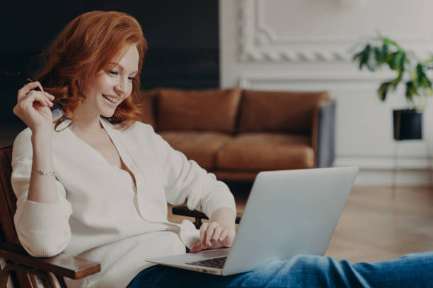 istock Skilled positive female student creats article for homework, sits in coworking space, has pleased expression, works on laptop computer, pose over home interior, prepares for writing course work 1466431412
