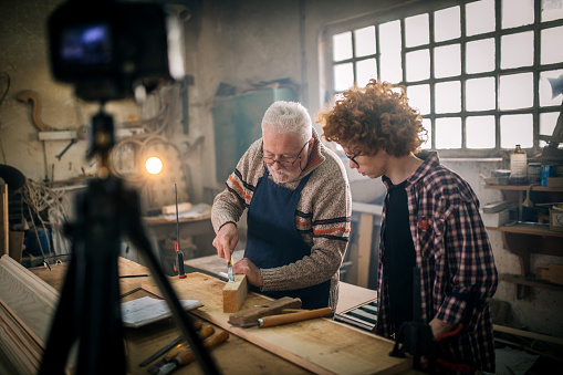 shot of senior craftsman and grandson recording a tutorial of him working on a project ind