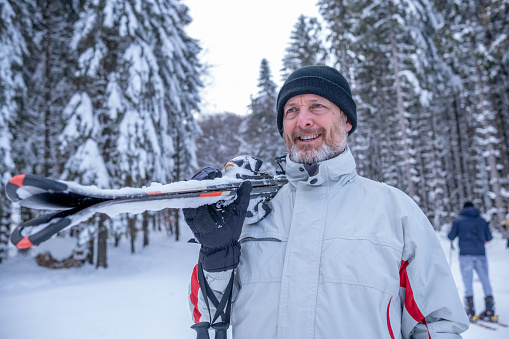 A cheerful man in a red-white ski outfit, black hat, and gloves with a ski on his shoulder on a ski slope.