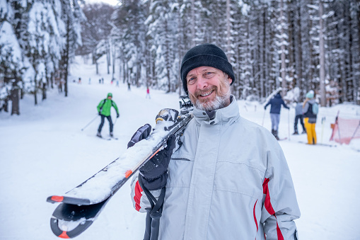 A cheerful man in a red-white ski outfit, black hat, and gloves with a ski on his shoulder is looking at the camera.