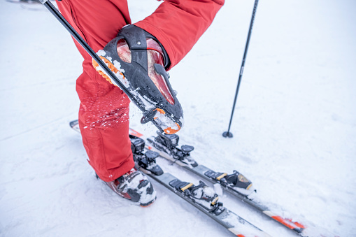 A man in red ski gear clears his ski boots of snow with a pole before attaching them to his skis.