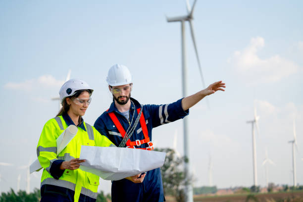istock Caucasian engineer man and woman discuss together using drawing paper and stay in front of row of windmill or wind turbine with blue sky and man also point forward. 1466437689
