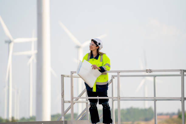 istock Engineer or technician woman worker hold drawing paper and stand on base of windmill or wind turbine and look to left side and stay in front of windmill cluster in background with blue sky. 1466437964