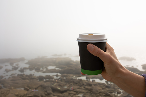 The girl's hand holds a cardboard cup for takeaway coffee in nature overlooking the ocean and heavy fog. Beautiful background, there is free space for the inscription.