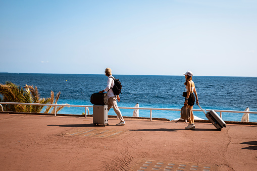 Young tourist couple with travel suitcases and bags walking on pedestrian walkway street at waterfront near blue sea water in Nice, France