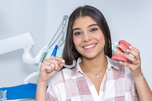 Delighted Hispanic female teenaged with long dark hair in casual clothes smiling and looking at camera while showing denture and transparent retainer during appointment at dental clinic