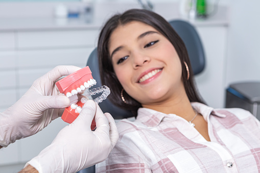 Crop unrecognizable dentist in latex gloves showing teeth mold and transparent silicone aligner to smiling Hispanic female teenager during appointment at dental clinic