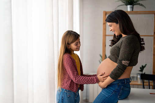 Beautiful happy pregnant woman spending time at home with her lovely daughter with long hair, bonding and cherishing precious moments. Sweet girl tenderly touching mother's belly, trying to feel the baby.