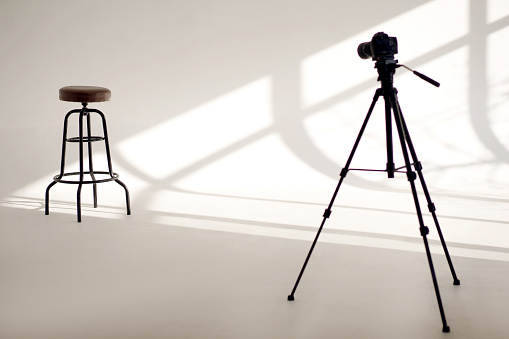 A white photo studio with sun shadows from the windows and one high chair and a video camera on a tripod. Professional photography with bar stool for model and white space with shadows