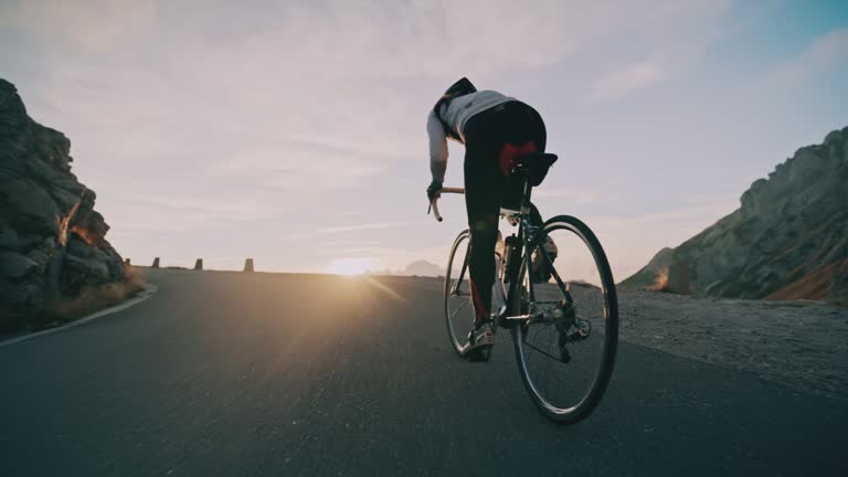 Tracking shot cyclist cycling uphill on road in sunrise mountains