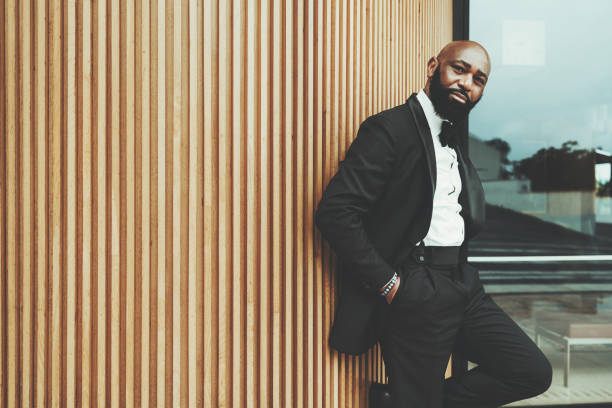 Black man leaning against wall A dandy black man with a bald head, wearing a black tuxedo, bow tie, and a white shirt; Hands in his pockets, and the other hand rests his body on a wall made of wood stakes creating an elegant design dinner jacket stock pictures, royalty-free photos & images