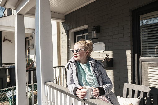 Millennial woman enjoying sunny day in front of the home and drinking coffee. She is dressed in casual clothes with winter jacket and sun glasses. Exterior of  private home in the city during sunny winter day.