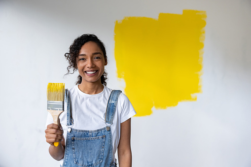 Happy African American woman remodeling her house and painting a wall yellow while looking at the camera smiling - home improvement concepts