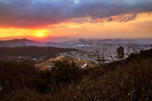Vista aérea de una ciudad detrás de las montañas en el atardecer, sol en el horizonte y cielo rojo con nubes moradas, bosque en las montañas, vista de Tultitlán, Tlalnepantla, estado de México y Ciudad de México, sierra de Guadalupe