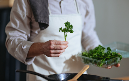 Unrecognizable chef in apron making a healthy meal with parsley while standing in the kitchen.