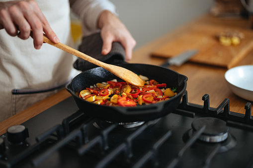 High angle view of an anonymous chef in apron standing at the gas stove and stirring vegetables in the frying pan.