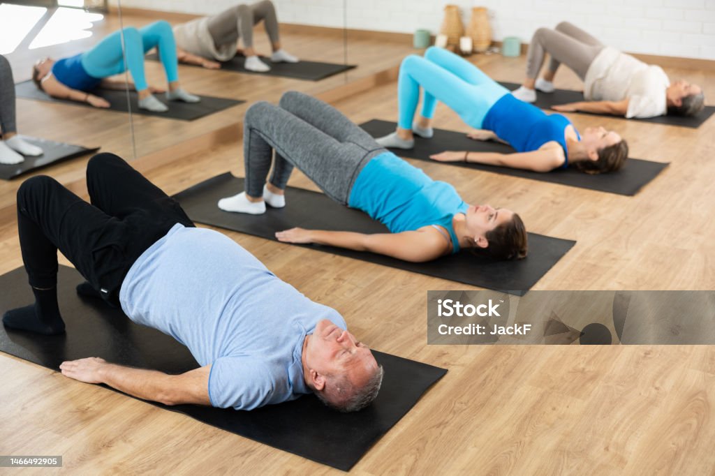 Aged man performing glute bridge in fitness studio Focused aged man exercising with group of people in fitness studio, performing glute bridge to strengthen core muscles, to tone glutes and hamstrings Exercising Stock Photo