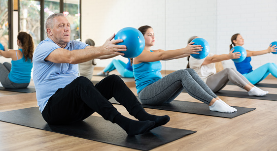 Diligent elderly man practicing pilates with ball in exercise room during pilates classes. Persons doing pilates in fitness hall.
